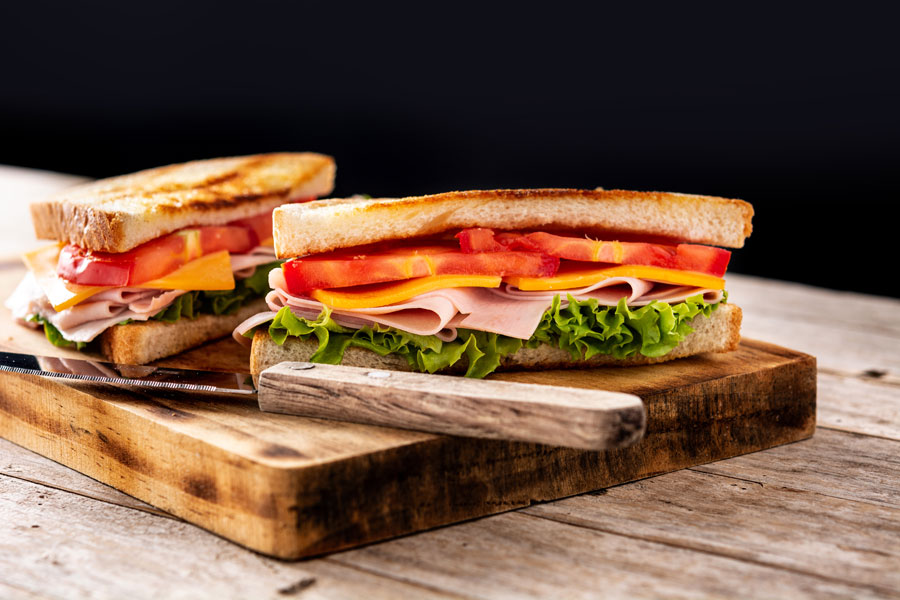 Fotografía de sandwich con lechuga, tomate, queso y jamón sobre tabla de madera.