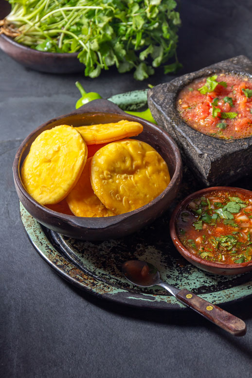 Fotografía de sopaipillas acompañadas de un mortero y cueco con pebre sobre una bandeja rústica sobre una mesa.